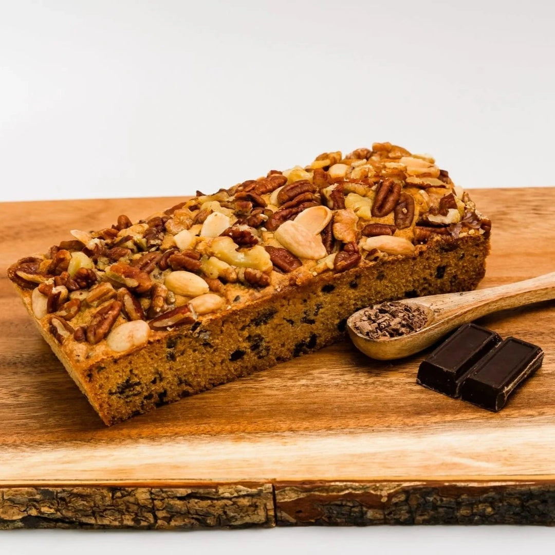 Loaf of bread with nuts on a wooden cutting board with chocolate pieces and a spoonful of something.