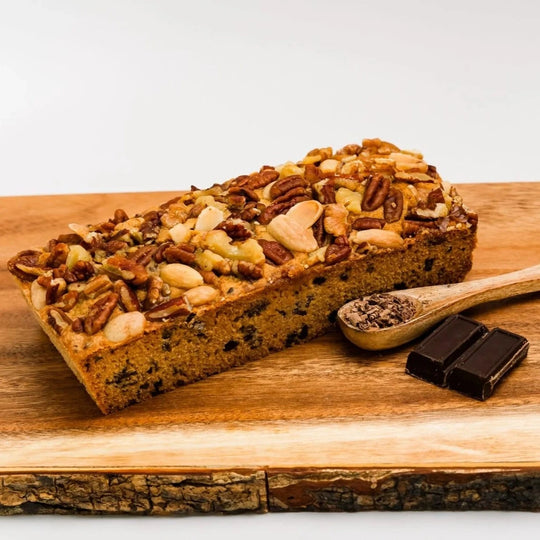 Loaf of bread with nuts on a wooden cutting board with chocolate pieces and a spoonful of something.