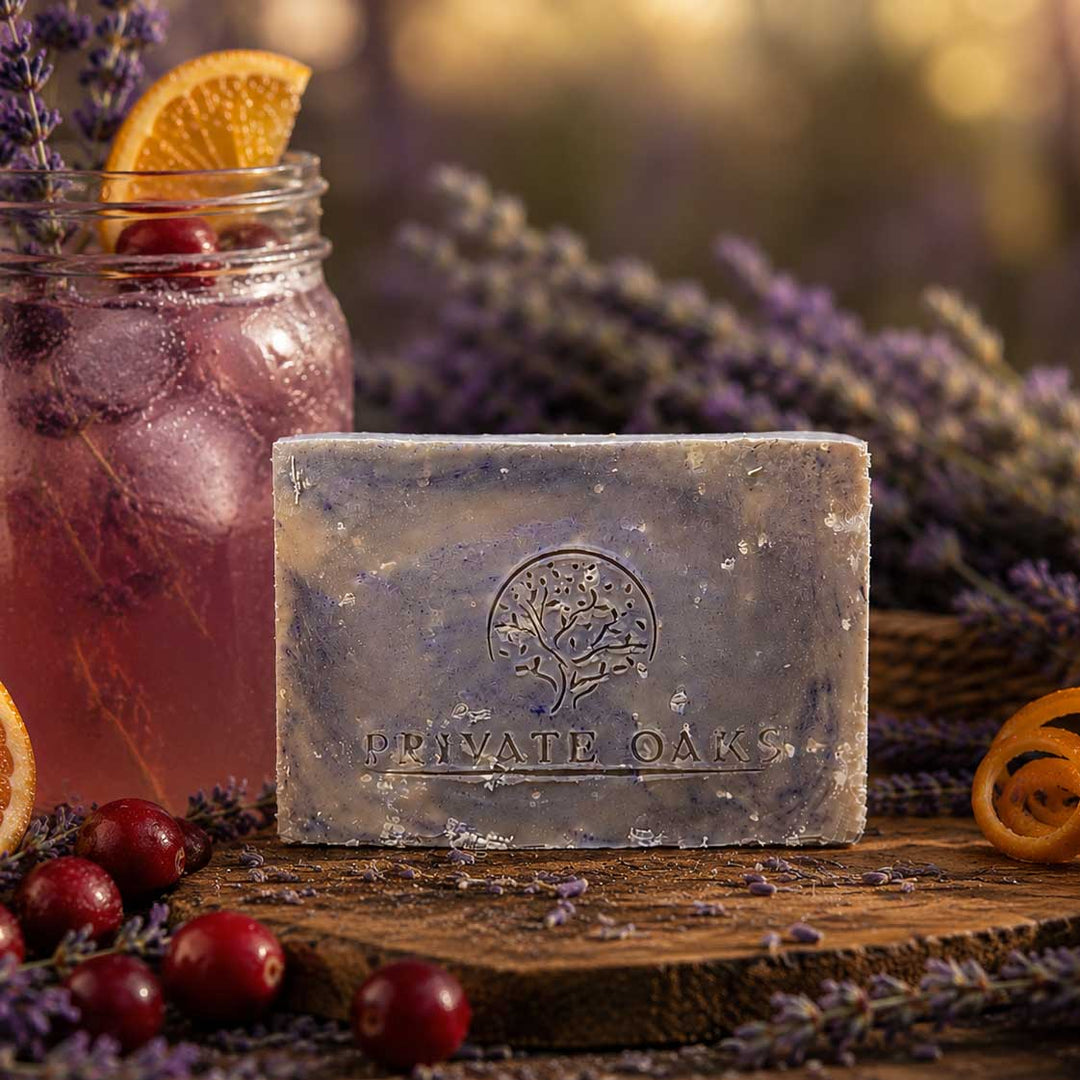 A rectangular bar of Private Oak soap with a tree logo embossed on the front, resting on a rustic wooden board surrounded by lavender, cranberries, and an orange slice. In the background, a glass jar holds a pink iced drink with an orange slice, set against a warm, softly blurred bokeh background.