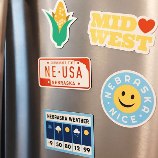 A collection of magnets with Midwestern themes displayed on a stainless steel surface, including a smiling face, corn, and state-related designs.