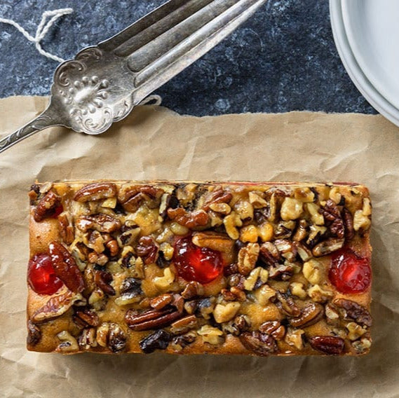 Rectangular fruitcake with nuts and cherries on brown paper with a fork and plates in the background.
