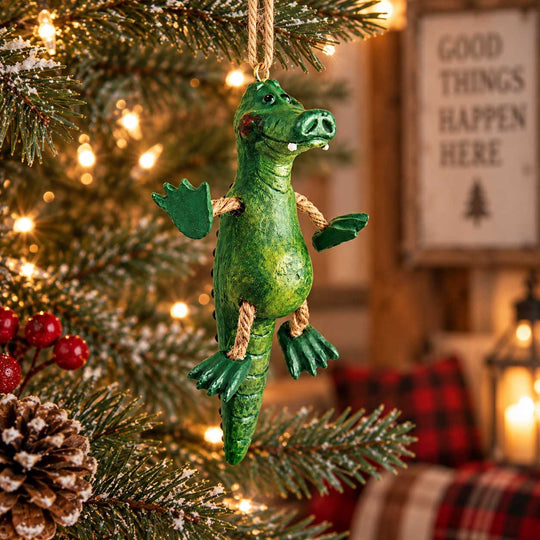 A green alligator Christmas tree ornament hanging from a string on a decorated Christmas tree, with warm lights and a cozy indoor background.