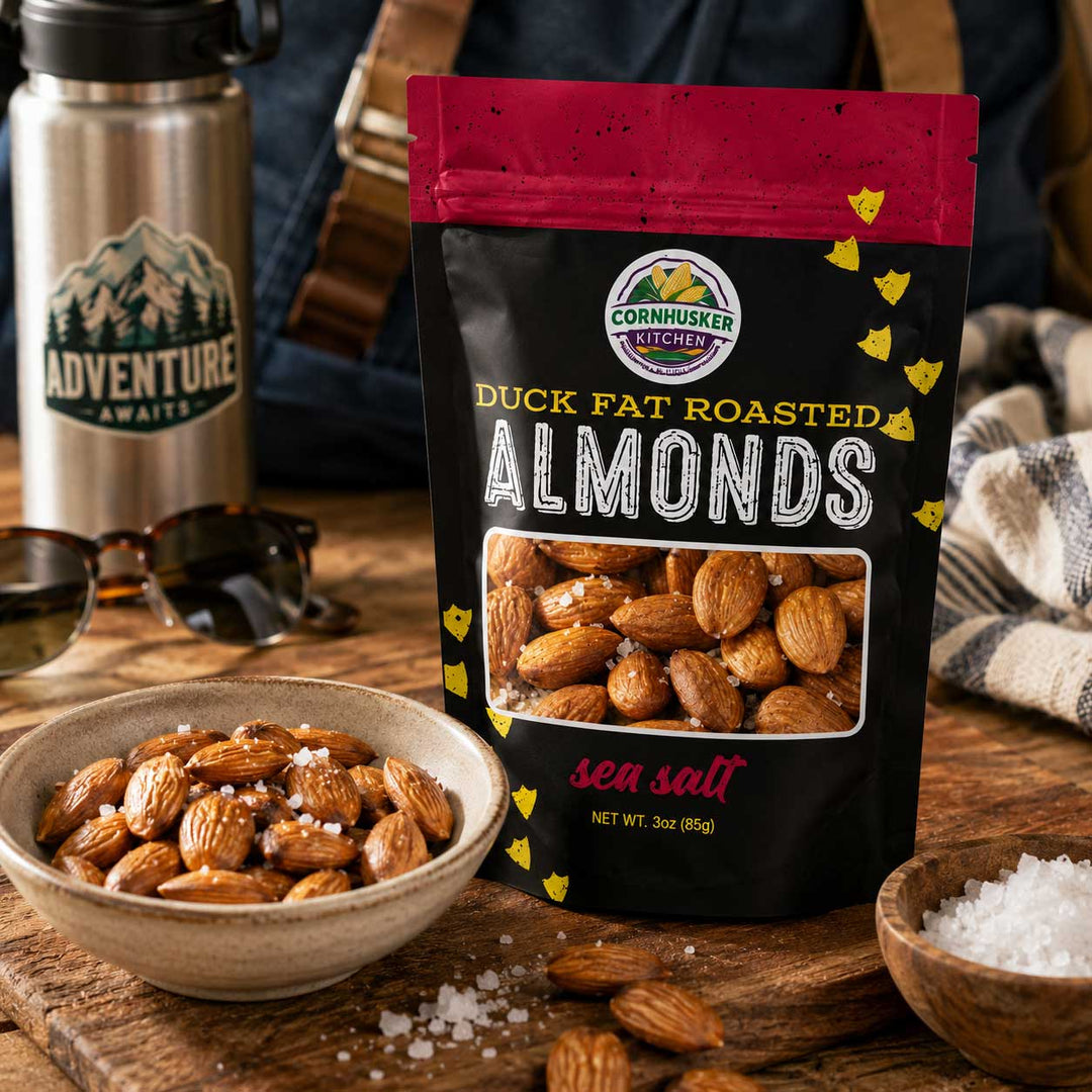 A bag of Cornhusker Kitchen Duck Fat Roasted Almonds with sea salt is displayed on a rustic wooden table next to a bowl of the almonds, a ceramic bowl of coarse salt, and a stainless steel travel tumbler with an Adventure Awaits logo.