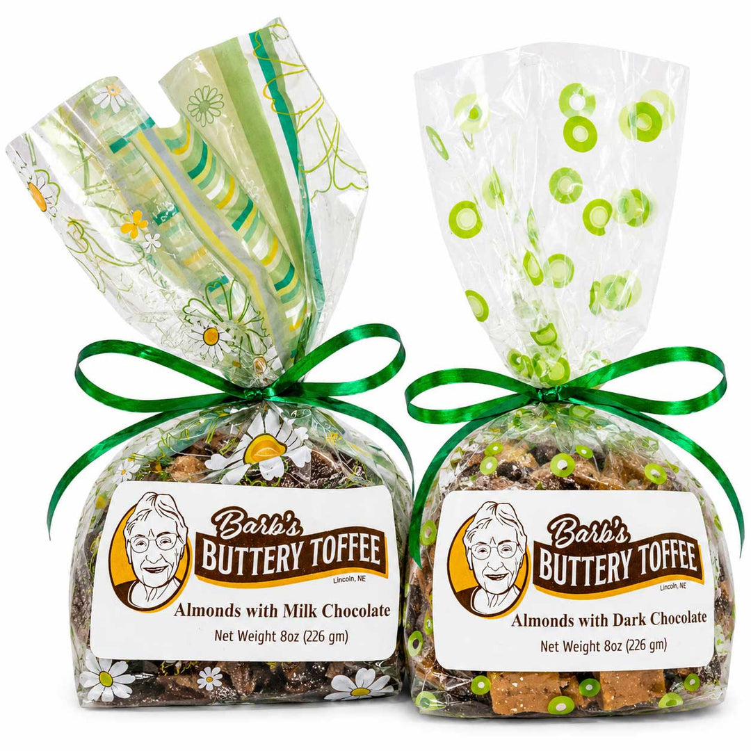 Two bags of Barb's Buttery Toffee, one with milk chocolate and one with dark chocolate, wrapped in clear cellophane with green accents and tied with green ribbons. The front of each bag features a white label with the brand logo and product name.