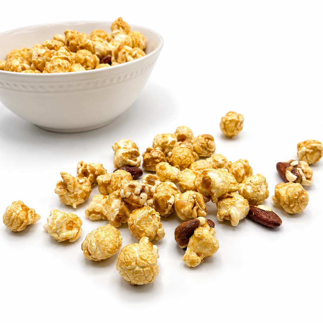 Caramel popcorn mixed with almonds scattered on a white surface, with a white bowl filled with more popcorn in the background.