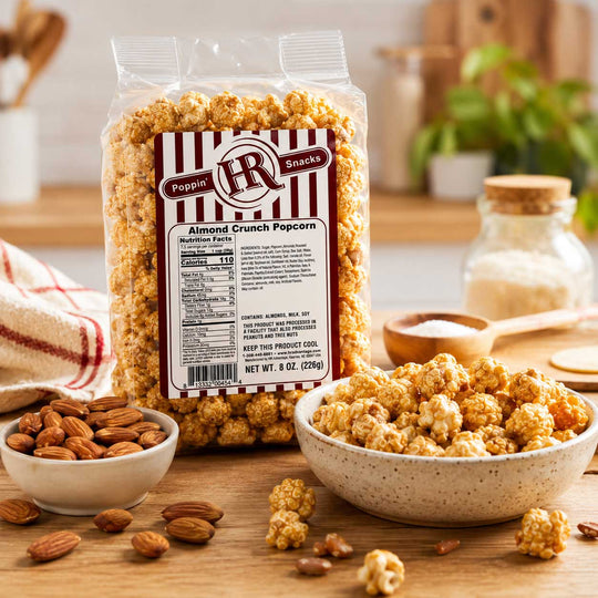 A bag of Poppin' Popcorns Almond Crunch Popcorn stands on a wooden table, surrounded by bowls of the popcorn, whole almonds, and a small jar in a warm kitchen setting.
