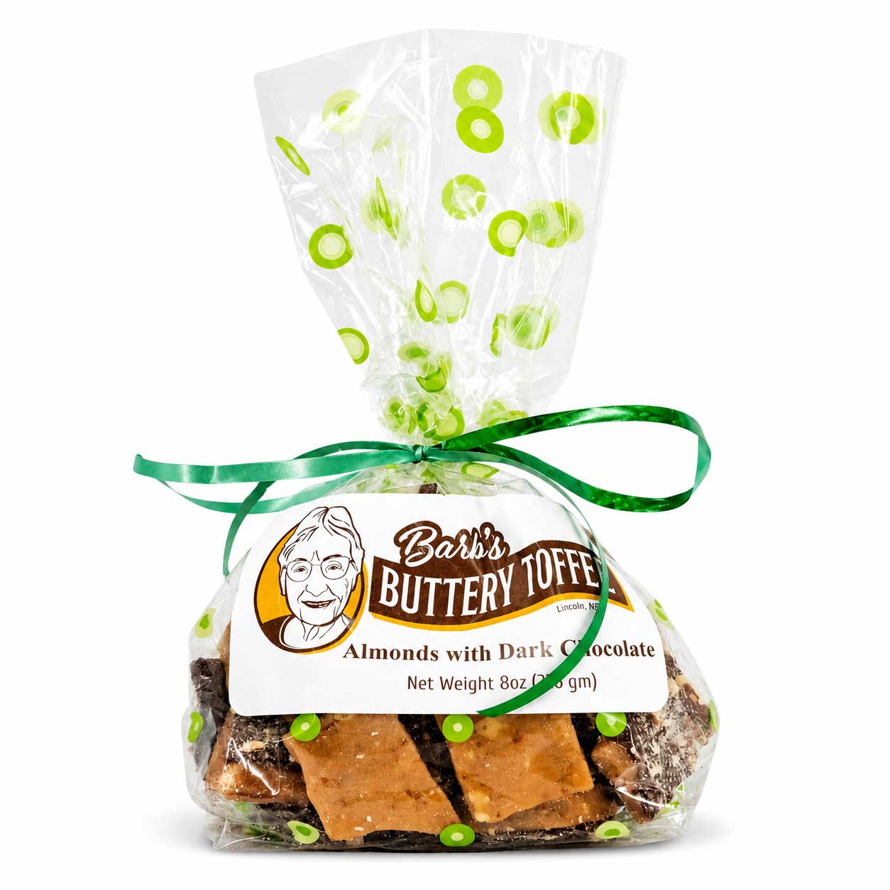 A clear cellophane bag of Barb's Buttery Toffee filled with almonds and dark chocolate pieces, tied with a green ribbon and decorated with green circular accents on the clear top seal, against a white background.