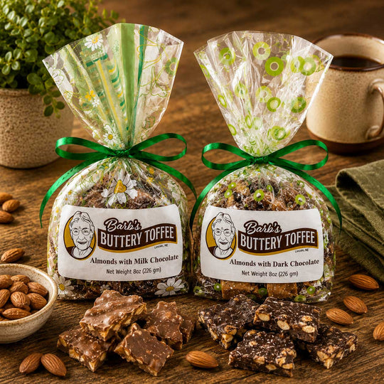 Two bags of Barb's Buttery Toffee, one with milk chocolate and one with dark chocolate, tied with green ribbons and displayed on a wooden table with loose almonds and chocolate pieces.