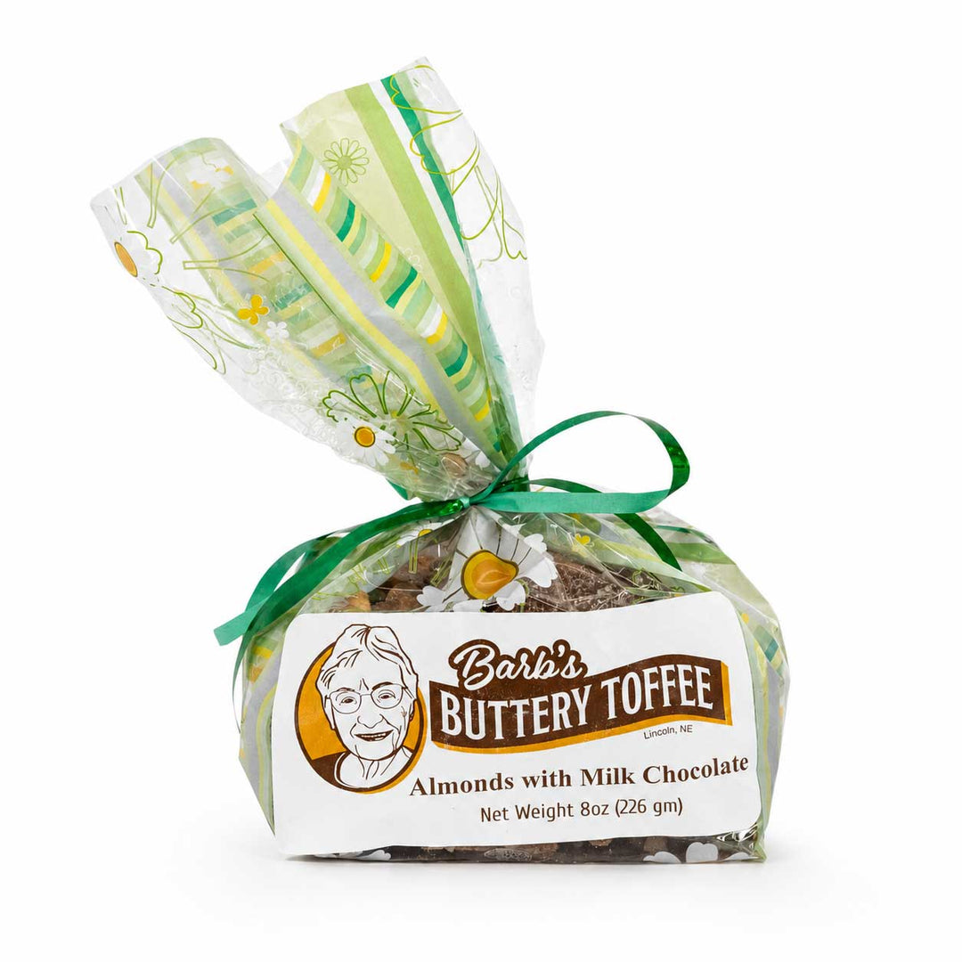 A packaged bar of butterscotch taffy wrapped in clear cellophane with green floral patterns, tied with a green ribbon, and featuring a white label that reads 'Barb's Buttery Toffee' on a white background.