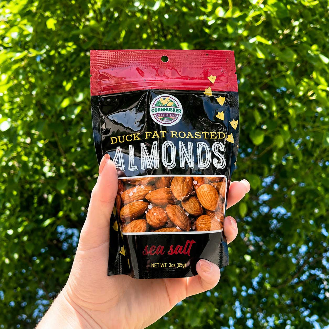 A hand holds a bag of Cornishions Duck Fat Roasted Almonds with sea salt against a background of green, sunlit foliage.