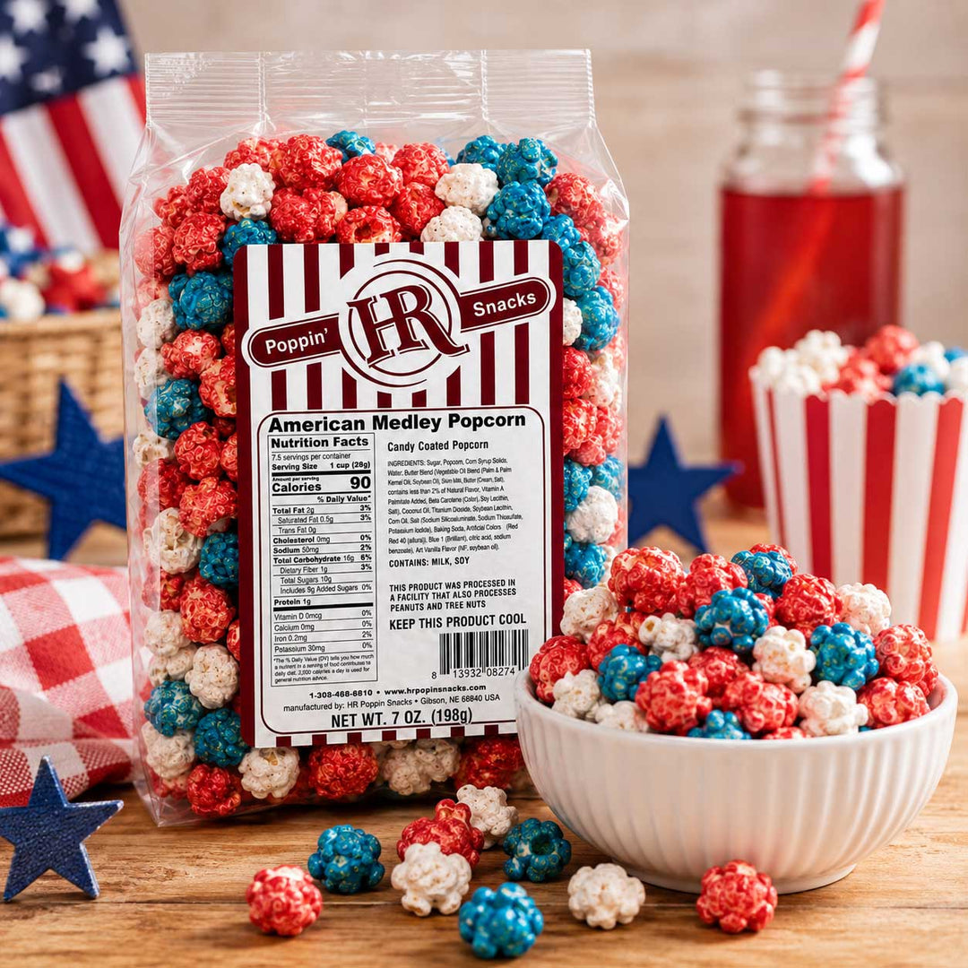 A bag of Poppin' IR American Medley Popcorn stands on a wooden surface with a white bowl, scattered popcorn pieces, a red-and-white striped cup, and patriotic props in the background. The product packaging is clear with a large front label and the brand logo visible.