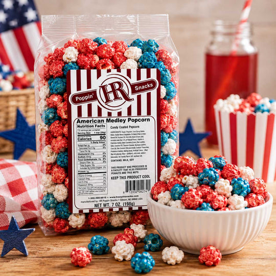 A bag of Poppin' IR American Medley Popcorn stands on a wooden surface with a white bowl, scattered popcorn pieces, a red-and-white striped cup, and patriotic props in the background. The product packaging is clear with a large front label and the brand logo visible.