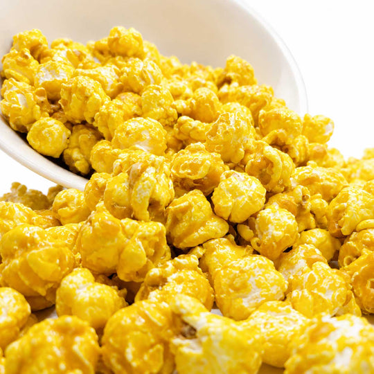 Close-up of bright yellow popcorn coated in a glossy glaze, spilling from a white bowl onto a white surface.