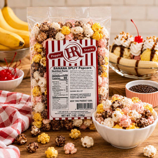 A 7 oz bag of Poppin' HR Banana Split Popcorn surrounded by bowls of the popcorn, banana slices, and dessert toppings on a wooden table.
