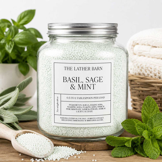 A glass jar candle filled with pale mint-green granules, labeled 'THE LATHER BARN BASIL, SAGE & MINT,' styled on a wooden surface with a spoon of the granules, fresh herbs, and a folded cream textile in the background.