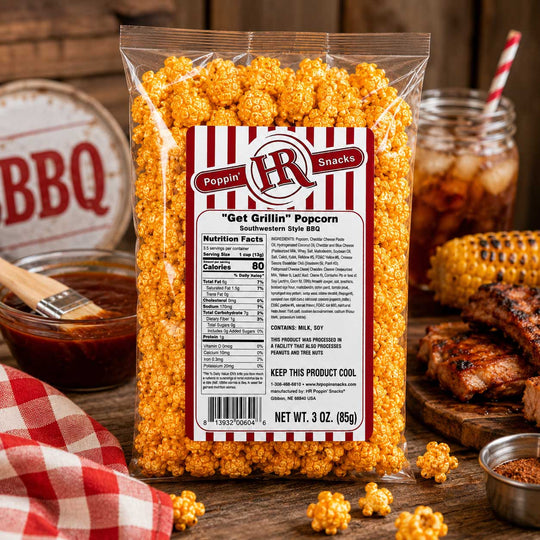 A 3 oz bag of Poppin' LR Snacks 'Get Grillin'' Popcorn (Southwestern Spice Blt) standing on a rustic wooden table surrounded by food props like grilled meat, a jar of sauce, and a red-and-white checkered cloth.