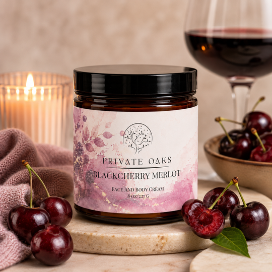 A jar of Private Oaks Blackcherry Merlot Face and Body Cream sits on a marble board surrounded by fresh cherries, a lit candle, and a glass of red wine in a warm, rustic setting.