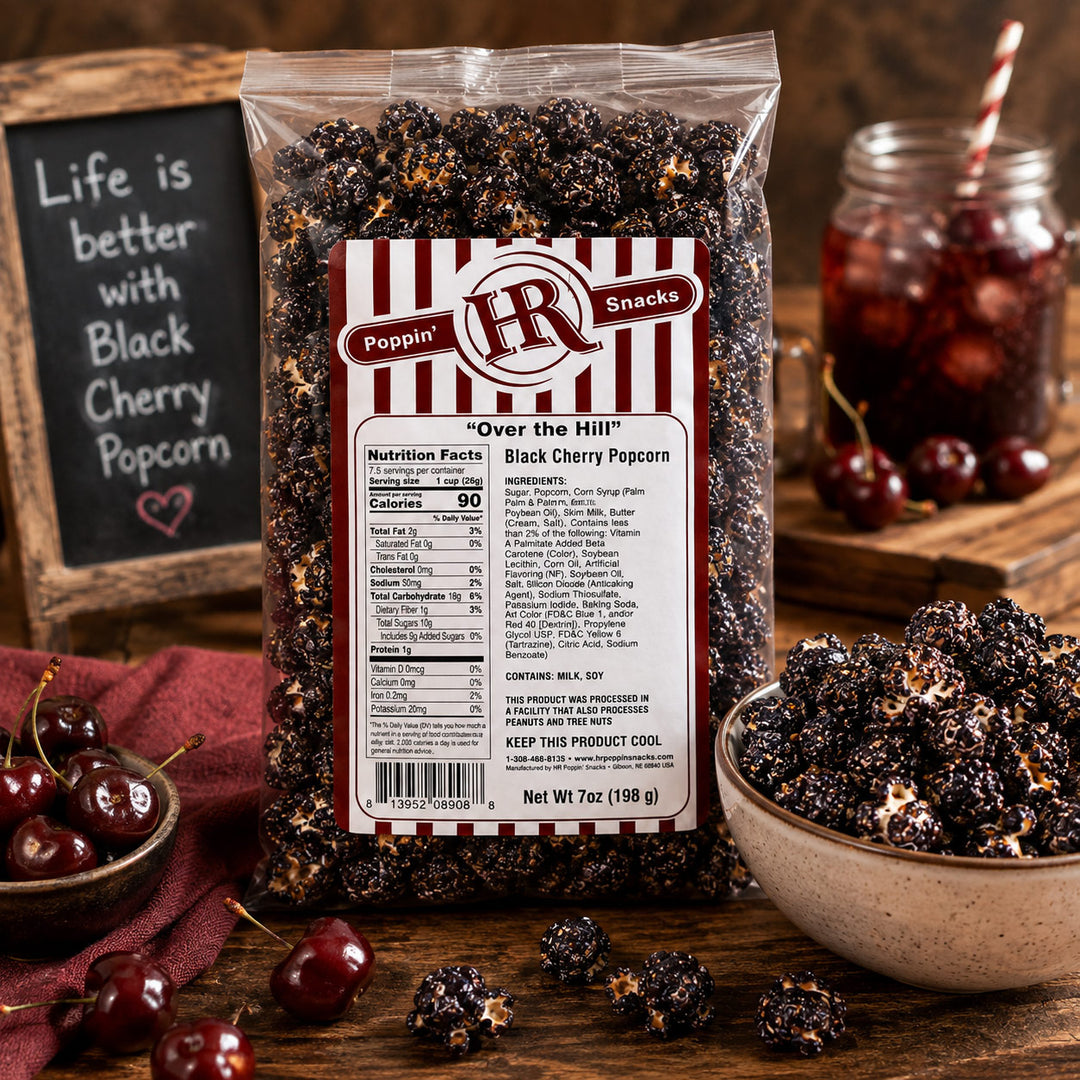 A bag of Poppin' R R Snacks 'Over the Hill' Black Cherry Popcorn is displayed on a rustic wooden table with bowls of the popcorn, fresh cherries, and a glass of cherries in the background, alongside a chalkboard sign.