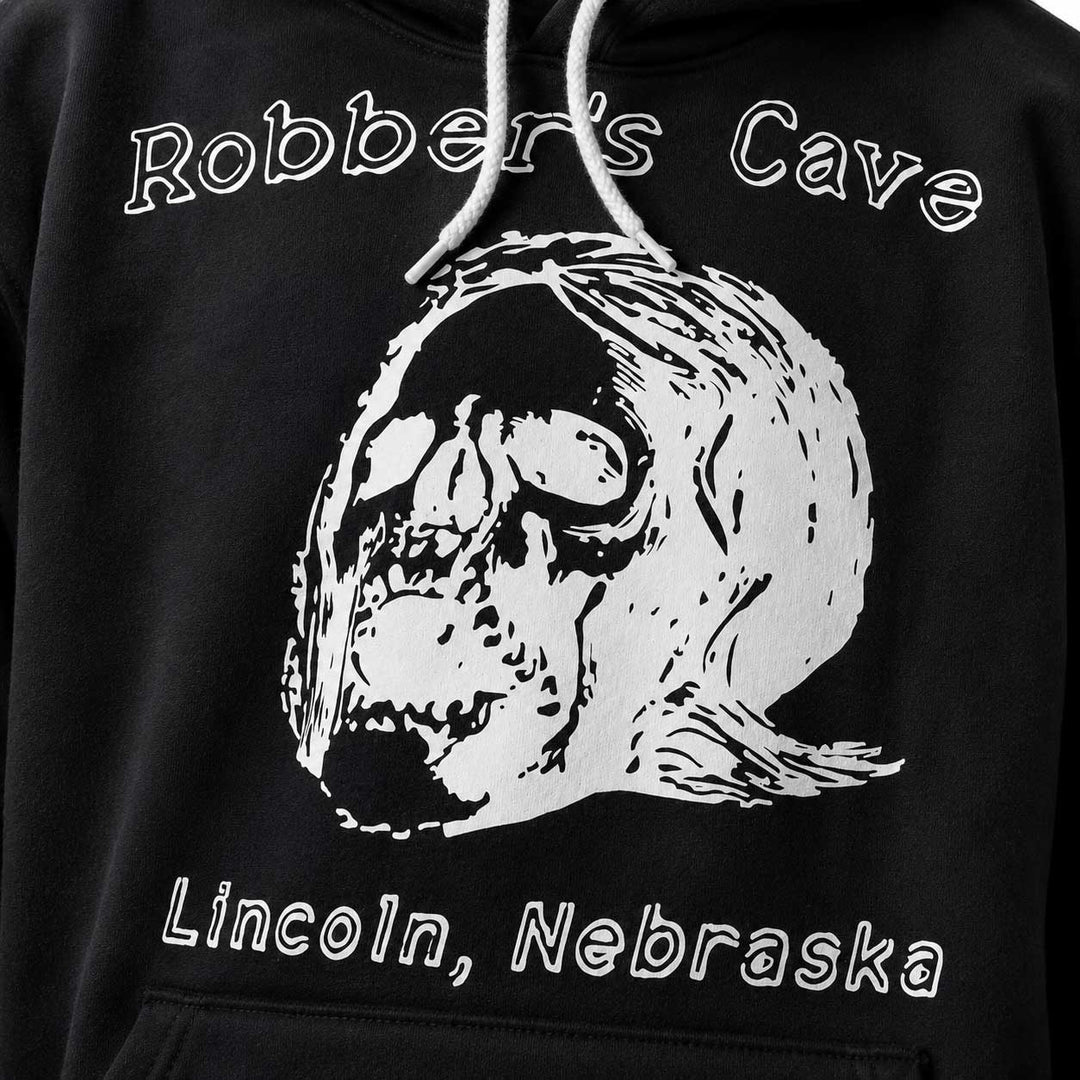 Close-up of a black hoodie featuring a large white graphic of a skull wearing a bandana, with the text 'Robber's Cave' above and 'Lincoln, Nebraska' below.