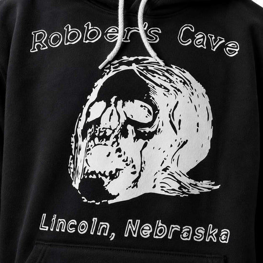 Close-up of a black hoodie featuring a large white graphic of a skull wearing a bandana, with the text 'Robber's Cave' above and 'Lincoln, Nebraska' below.