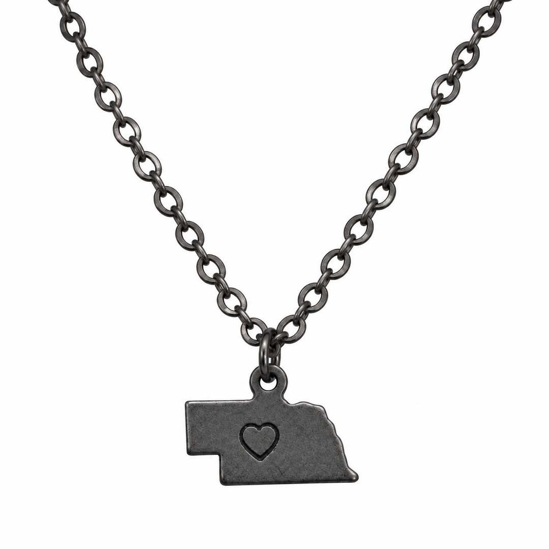 A dark gunmetal gray necklace featuring a pendant shaped like the state of Nebraska, with a small heart cutout in the center, displayed against a solid white background.