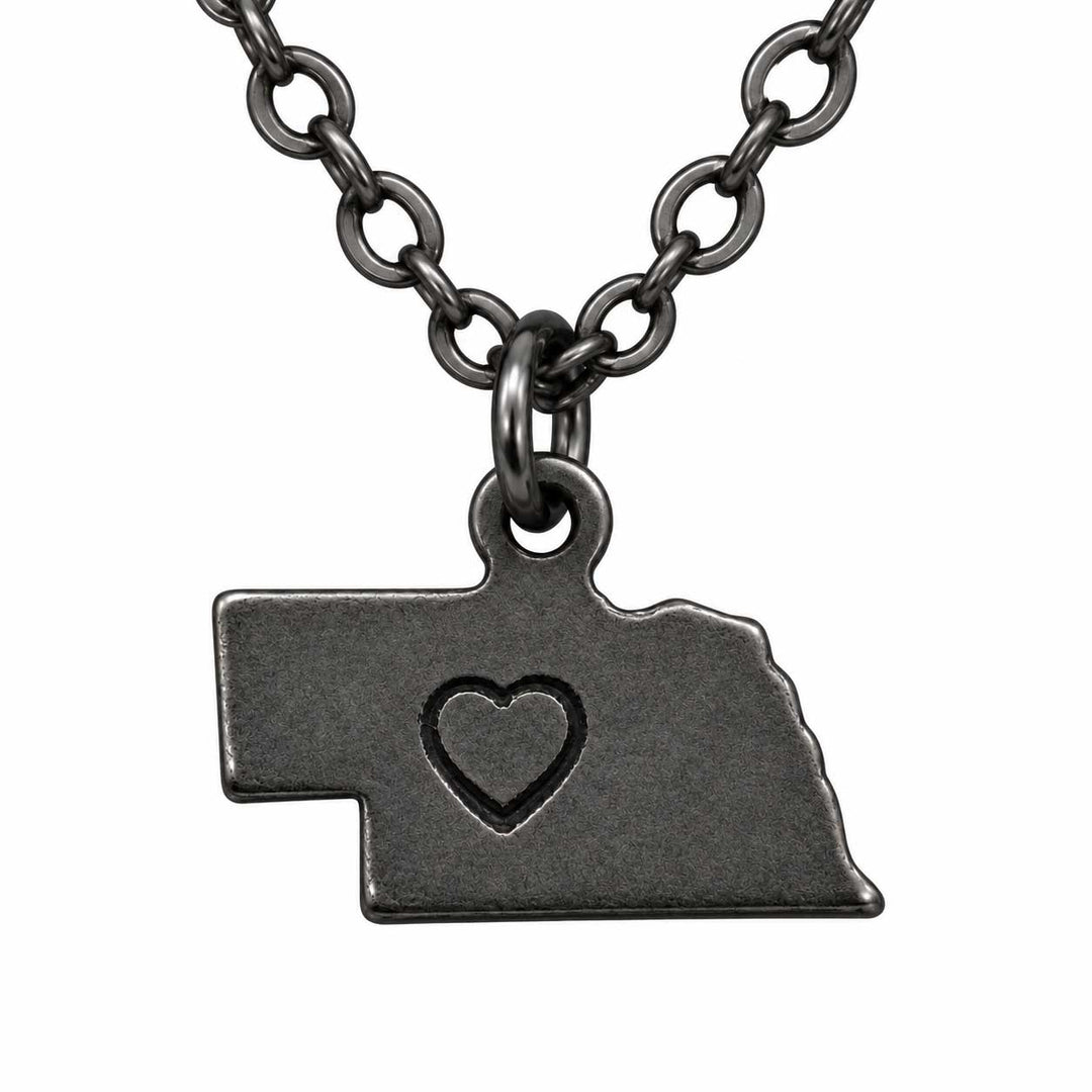 A dark gunmetal gray necklace featuring a pendant shaped like the state of Nebraska, with a small heart cutout in the center, displayed against a solid white background.