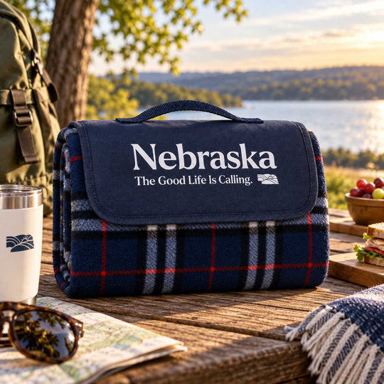 A navy blue picnic blanket with a plaid pattern in red, white, and gray, featuring the text 'Nebraska The Good Life Is Calling.' on the front flap.