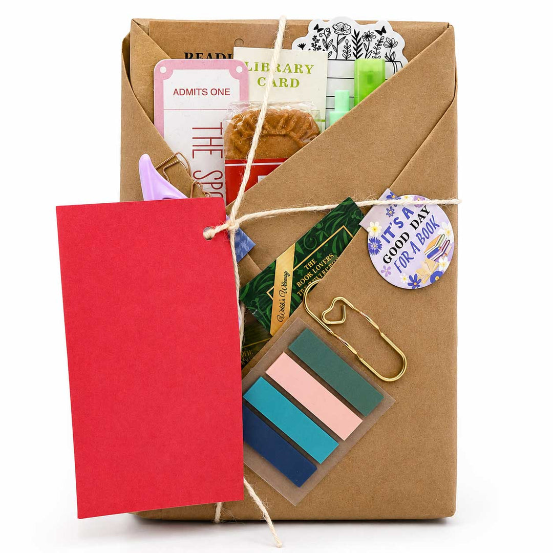 A brown kraft paper envelope-style book gift box tied with twine, filled with stationery and small accessories including a red tag, colorful sticky notes, cards, a gold paperclip, and a round sticker, on a white background.