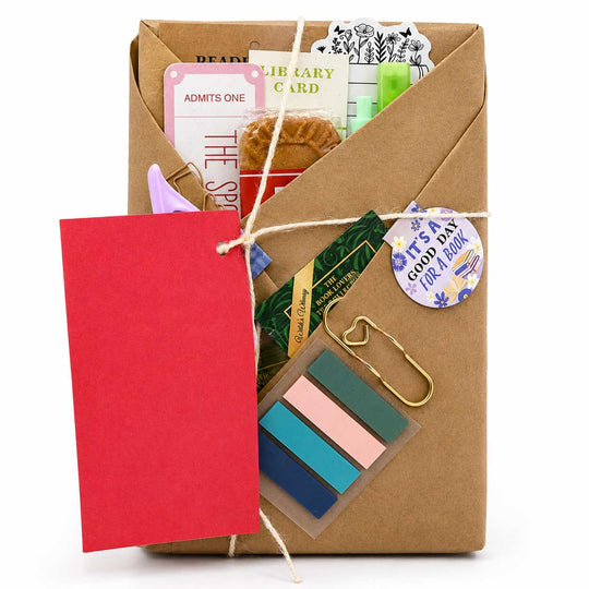 A brown kraft paper envelope-style book gift box tied with twine, filled with stationery and small accessories including a red tag, colorful sticky notes, cards, a gold paperclip, and a round sticker, on a white background.