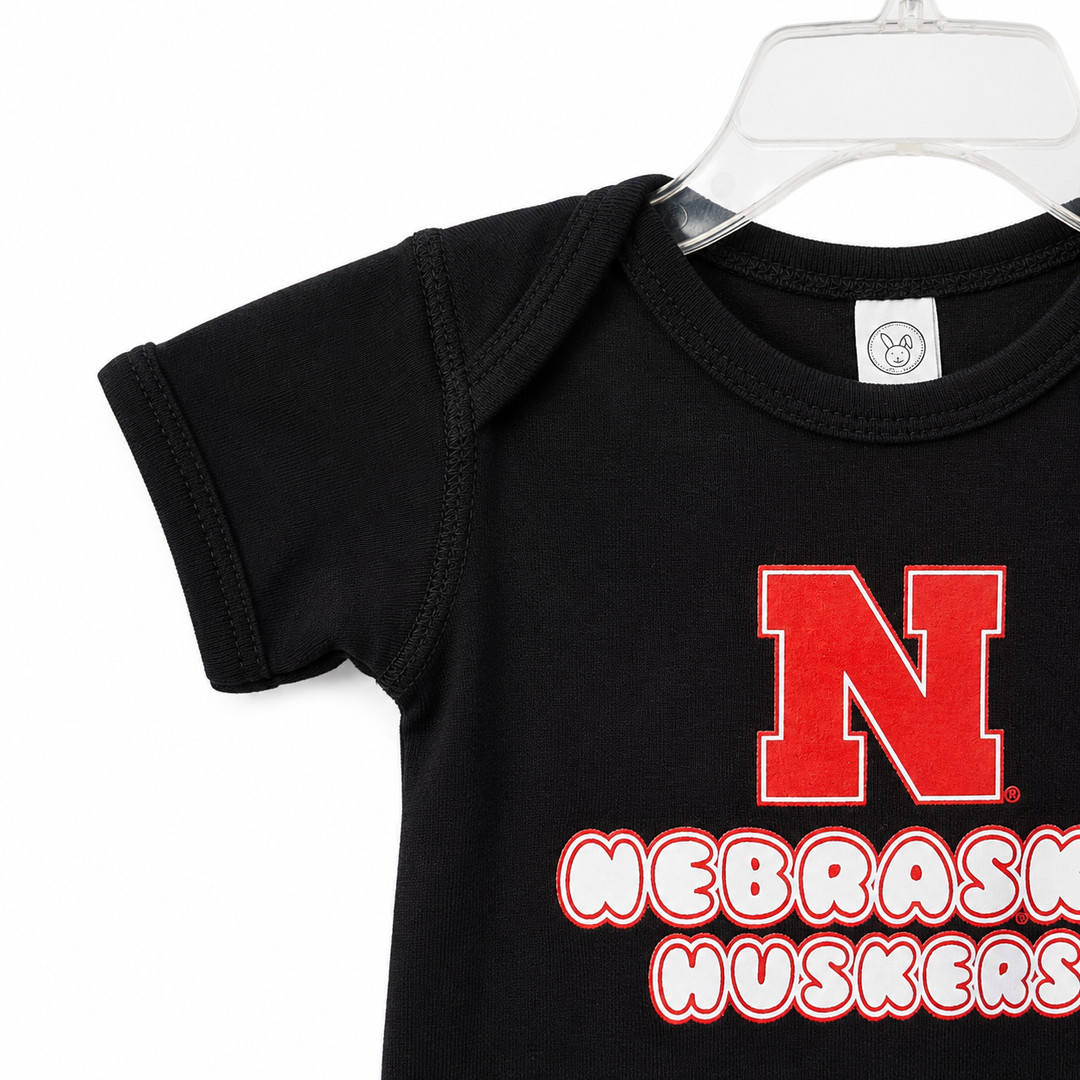 Black baby onesie with red and white 'Nebraska Huskers' logo on a hanger.