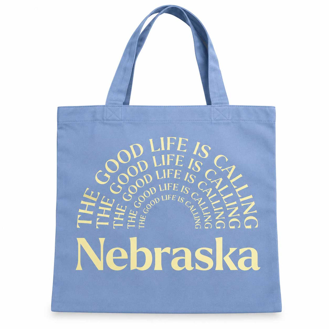 A light blue canvas tote bag with yellow text that reads 'THE GOOD LIFE IS CALLING Nebraska' in a circular design, shown against a plain white background.