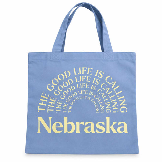 A light blue canvas tote bag with yellow text that reads 'THE GOOD LIFE IS CALLING Nebraska' in a circular design, shown against a plain white background.