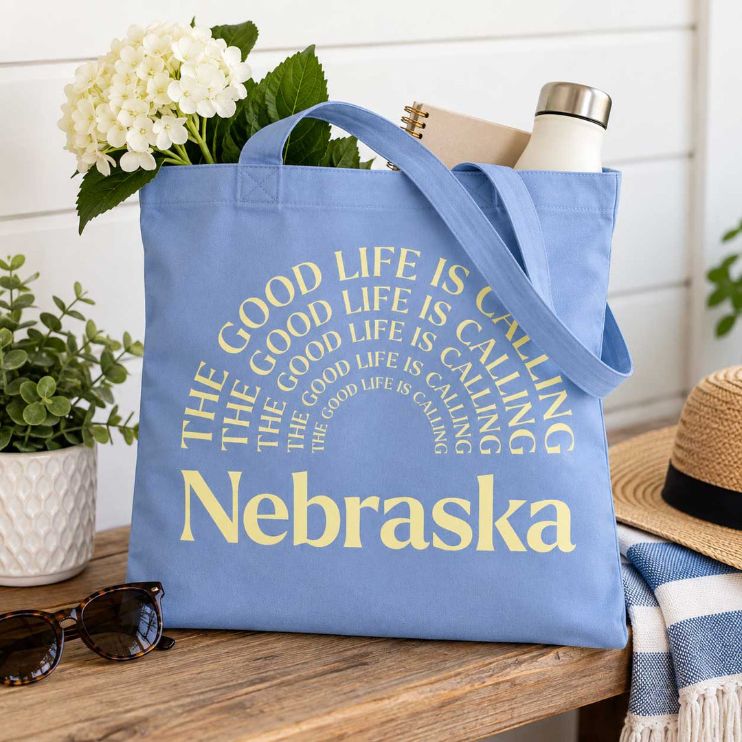 A light blue canvas tote bag with yellow text reading 'THE GOOD LIFE IS CALLING Nebraska' sits on a wooden table in a styled setting with flowers, sunglasses, a straw hat, and a blanket.