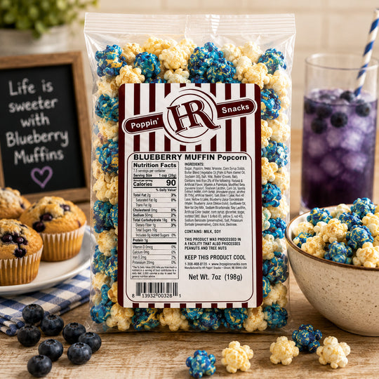 A 7 oz bag of Poppin' Snacks Blueberry Muffin Popcorn stands on a wooden table surrounded by blueberry muffins, a bowl of popcorn, blueberries, and a glass of purple drink. The bag has a clear plastic body with a large white and red striped label showing the brand 'Poppin' Snacks' and product details.