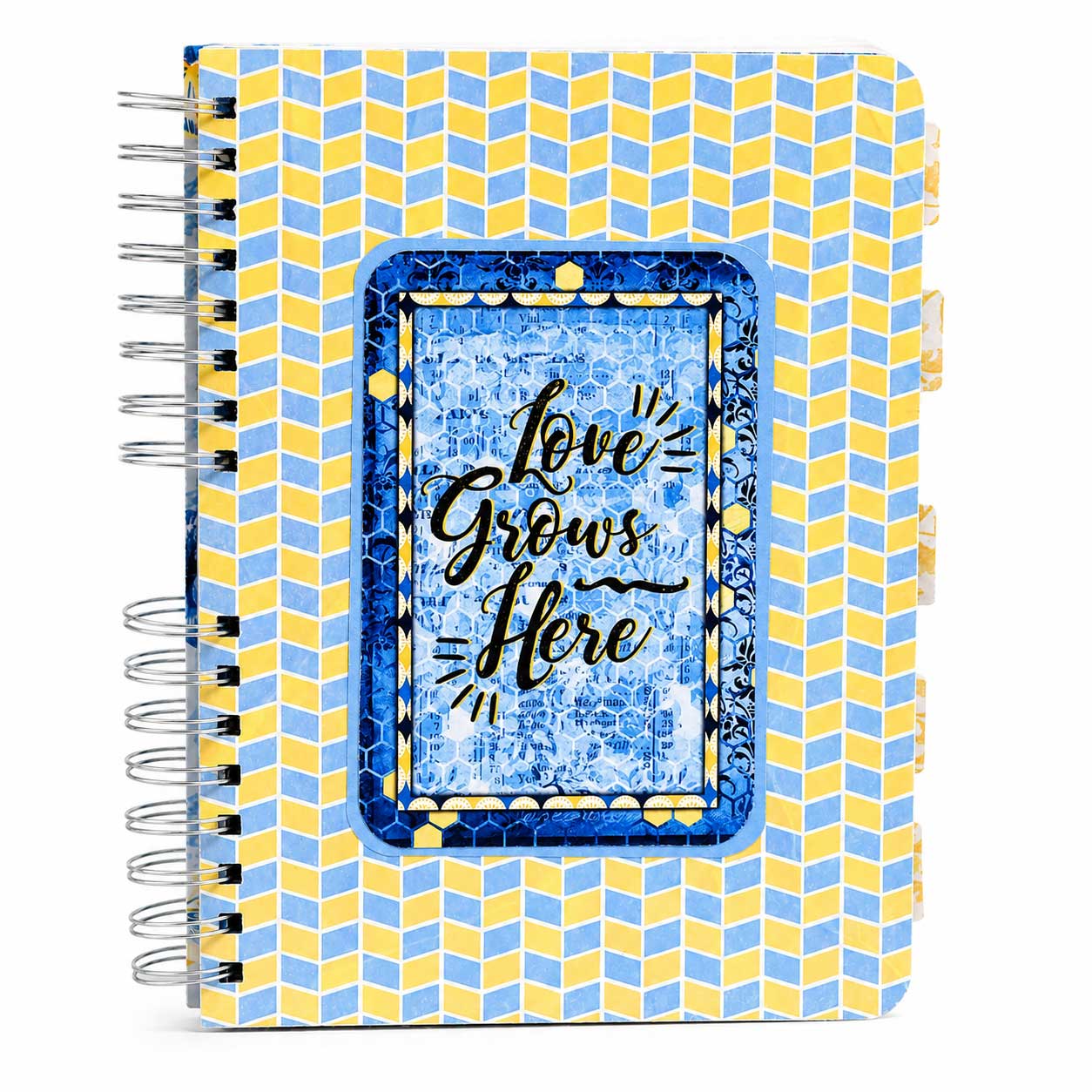 A spiral-bound notebook with a yellow and light blue geometric chevron pattern cover, featuring a central blue textured insert with the phrase 'Love Grows Here' in black script.