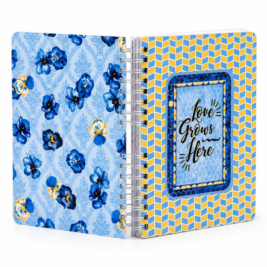 A spiral-bound notebook shown open, featuring a light blue floral pattern on the left cover and a yellow geometric pattern on the right cover. The right cover has a blue decorative frame with the words 'Love Grows Here' in black script.
