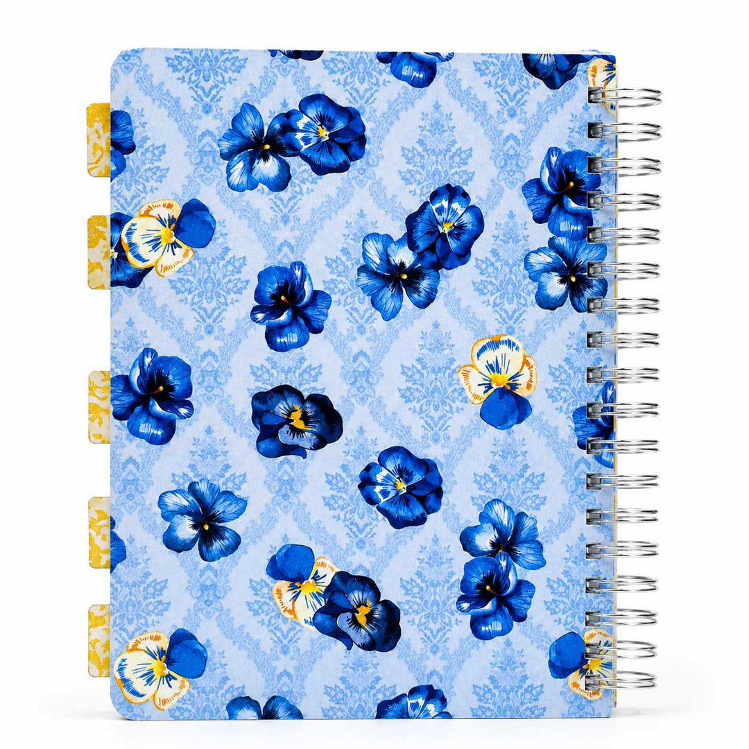 A spiral-bound planner or notebook with a light blue damask pattern cover decorated with blue and yellow pansy flowers. It features gold-toned page tabs on the left margin and a silver wire spiral binding on the right.
