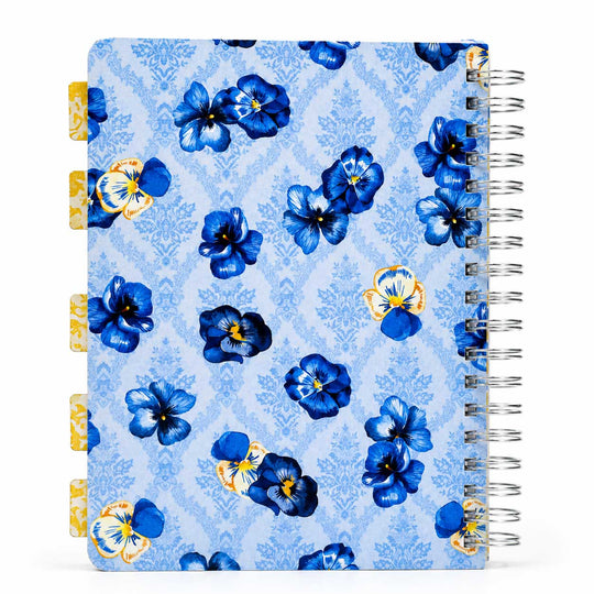 A spiral-bound planner or notebook with a light blue damask pattern cover decorated with blue and yellow pansy flowers. It features gold-toned page tabs on the left margin and a silver wire spiral binding on the right.