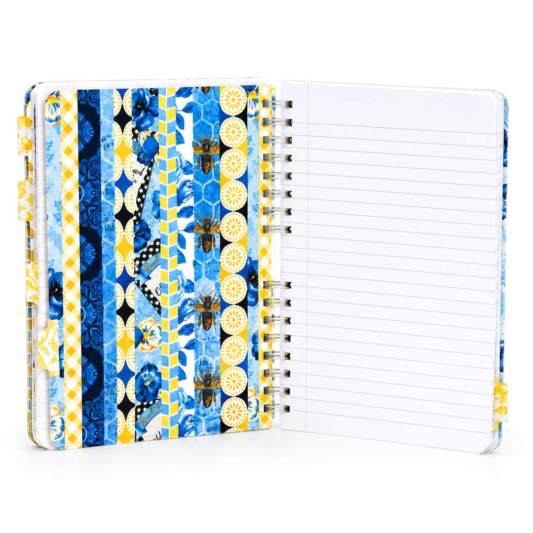 An open spiral-bound notebook with a decorative cover featuring blue, yellow, and white geometric patterns and floral motifs, next to blank lined pages.