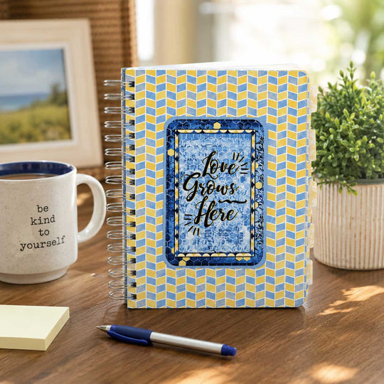 A spiral-bound planner or notebook with a blue and yellow chevron pattern and a central blue label reading 'love grows here,' styled on a wooden desk beside a mug, pen, sticky notes, and a plant.