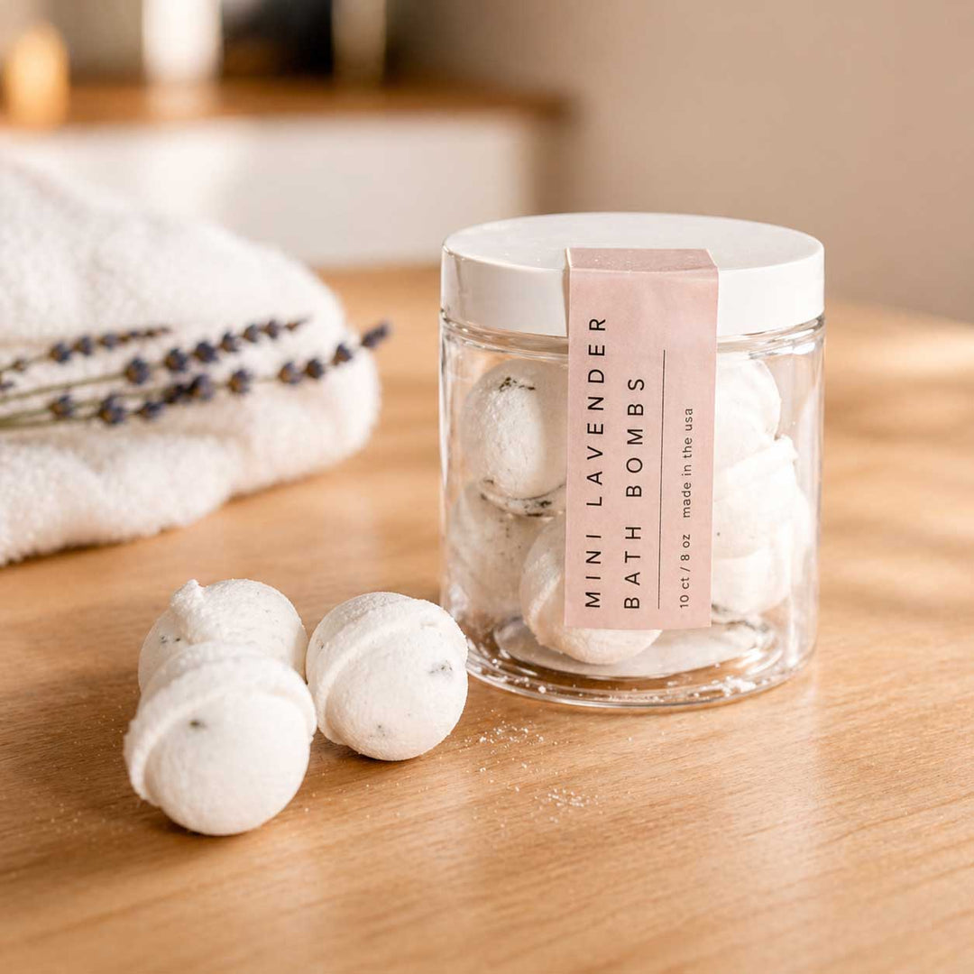 Jar of Mini Lavender Bath Bombs on a wooden surface with a towel and lavender flowers in the background.