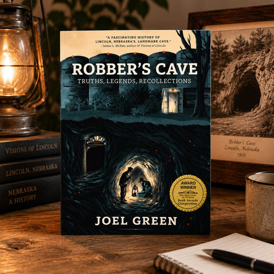 The book 'Robber's Cave' by Joel Green is displayed upright on a wooden table, surrounded by thematic props including a lit vintage lantern, a notebook, and another book in the background.