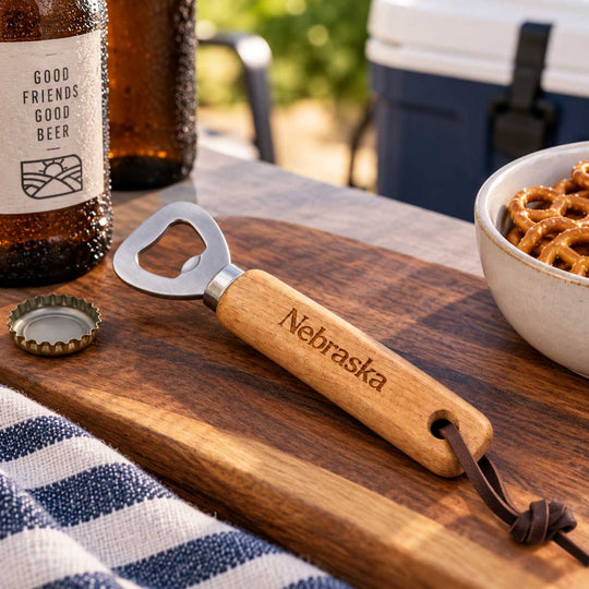 A wooden bottle opener engraved with the word 'Nebraska' rests on a wooden cutting board outdoors. A bottle cap lies beside it, with beer bottles, a bowl of pretzels, and a cooler visible in the blurred background.