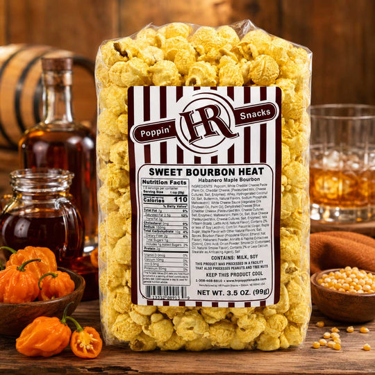 A clear plastic bag of Poppin' HR Snacks Sweet Bourbon Heat popcorn stands on a rustic wooden surface with bottles, glasses, and bowls of ingredients around it. The bag is filled with yellow-orange flavored popcorn and has a white label with black stripes and the Poppin' HR logo.