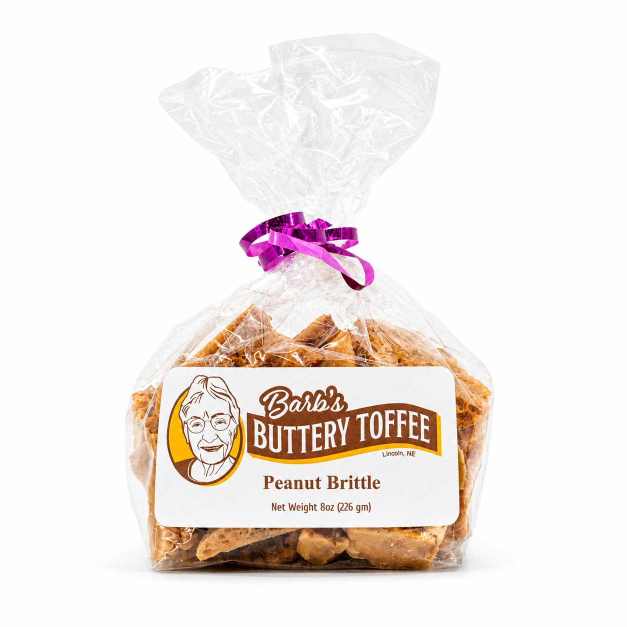 A clear plastic bag of Barb's Buttery Toffee Peanut Brittle tied with a purple ribbon, with a white and brown product label on a white background.