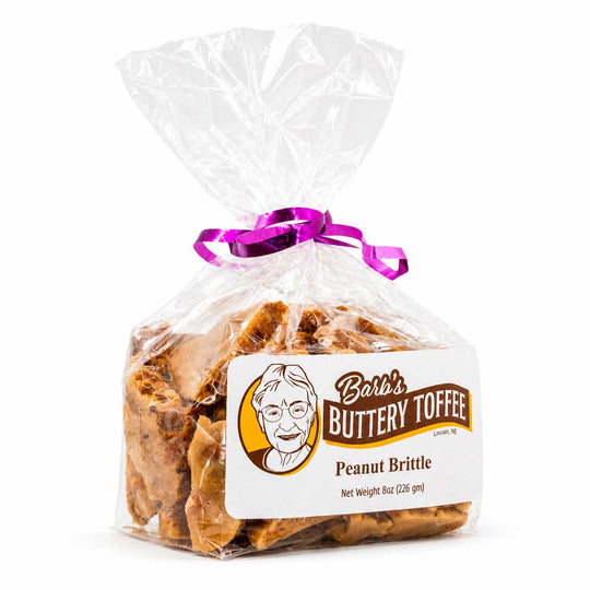 A clear plastic bag of Barb's Buttery Toffee Peanut Brittle tied with a purple ribbon, with a white and brown product label on a white background.