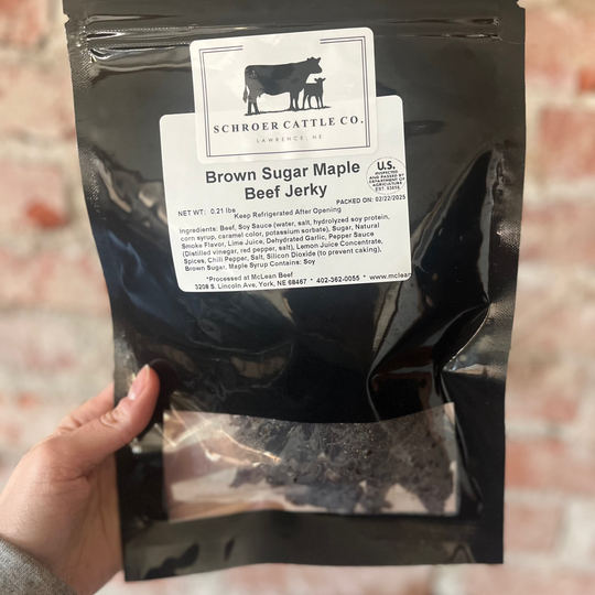 Black bag of Brown Sugar Maple Beef Jerky with Schrider Cattle Co. label held by a hand against a brick wall.