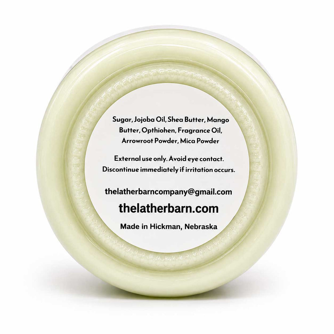 Top-down view of a round white jar lid with a white label containing product ingredients and the brand name 'thelatherbarn.com' on a white background.