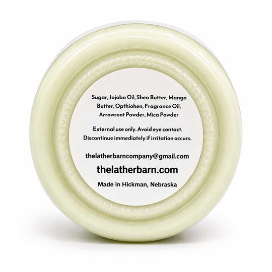 Top-down view of a round white jar lid with a white label containing product ingredients and the brand name 'thelatherbarn.com' on a white background.
