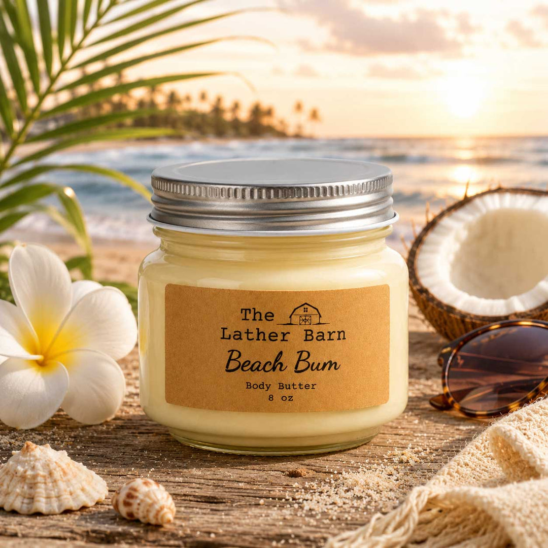 A jar candle labeled 'The Lather Barn Beach Bum' sits on a beach-themed surface with a sunset ocean background, surrounded by a white plumeria flower, a halved coconut, sunglasses, shells, and a woven cloth.